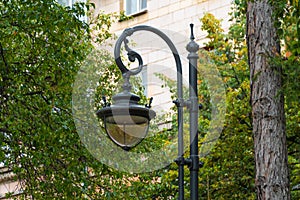 Old black cast iron lantern with round lampshade in park against background of green trees