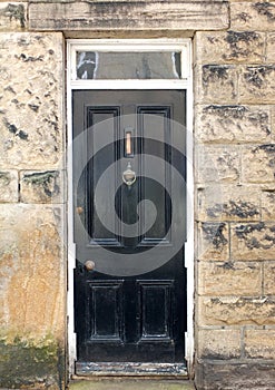 Old black painted wooden house door with brass letter box and knocker with a white frame surrounded by a stone wall
