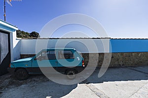 Old blue car parked ear a garage under the sunlight and a blue sky