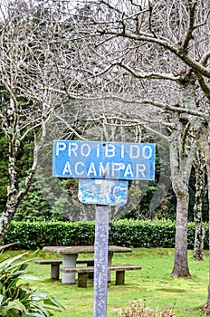 Old blue sign with text Proibido Acampar in Portuguese language. TRANSLATION: Camping forbidden. Park with green trees