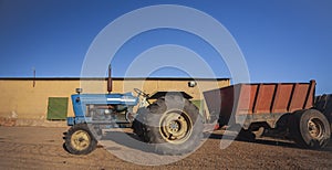 An old blue wheeled tractor with trailer stands in rural yard