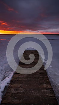 An old boardwalk pier on a large frozen lake with a dramatic evening sky and sunset crimson over the horizon