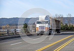 Old bonnet cab-over big rig semi truck with bulk trailer running on the road with protection fence on the side