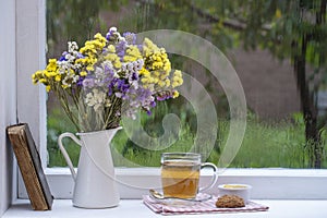 Old book, colorful bouquet of flowers and white cup of tea on background of window with raindrops at home at summer day