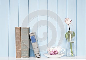 Old books, china cup and rose in the bottle on bookshelf