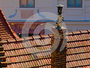Old brick and cast iron chimney above light brown clay tile roof ridge and blurred eclectic stucco facade background