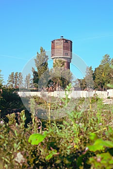 An old brick water tower with an outbuilding made of wooden boards at the top, behind a concrete fence, in the rays of the rising