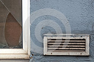 Old broken glass window on a blue house facade with a dirty ventilation grille