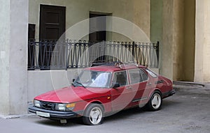 An old broken red car with a flat tire is parked against the wall of a house