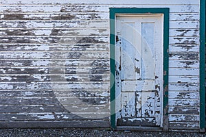 This old building has narrow wood slats with a wooden panel door. The colors are white and green & paint is worn away.