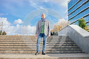 Old businessman in stylish scarf stand on paved stairs on blue sky background, from below view. Man walk outdoors.