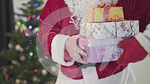 Old Caucasian man in Santa Claus costume giving presents to camera in front of the New Year tree. Man holding gift boxes