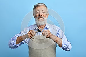 Old cheerful chef or waiter in gray apron, blue shirt holding fork, spoon