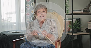 Old cheerful lady sitting on chair and knitting at her apartment. Front view of elderly woman looking surprised while