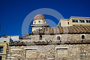 Old classic little church in earth tone natural stone with pigeons on terracotta roof tile with clear blue sky and modern building