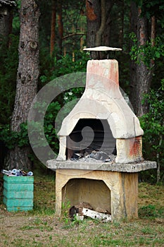 Old clay furnace with logs standing outdoors near the forest and full garbage bin