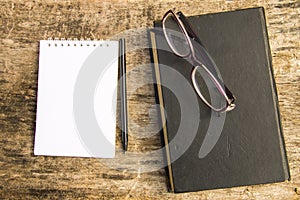 Old closed book, glasses, notepad and pen on wooden background