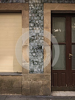 Old closed window with wooden shutter and a door with translucent glass in a granite stone wall and blue tiles