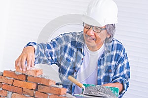 Old construction worker laying down brick wall with cement, wearing protected builder hard hat