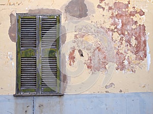 Old cracked house wall with patched cement painted in flaking peeling layers of grey yellow and red paint with a green wooden c