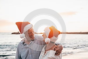 Old cute couple of mature persons enjoying and having fun together at the beach wearing christmas hats on holiday days. Hugged on