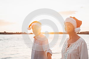 Old cute couple of mature persons enjoying and having fun together at the beach wearing christmas hats on holiday days. Walking on