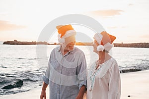 Old cute couple of mature persons enjoying and having fun together at the beach wearing christmas hats on holiday days. Walking on