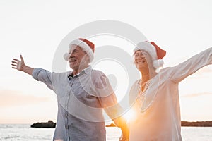 Old cute couple of mature persons enjoying and having fun together at the beach wearing christmas hats on holiday days. Walking on