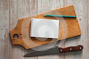 An old cutting board, a chef`s knife, a Notepad for writing recipes and a pencil on a light wooden background.