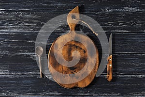 An old cutting board with a spoon and a knife on a rustic black background.