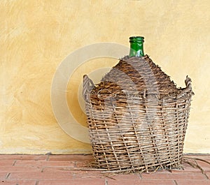 Old demijohn aka carboy for wine, wicker straw basket. by house. Y