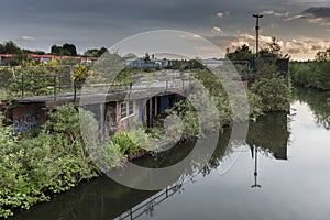 An old, derelict, building, beside a calm canal, overgrown with foliage, set in a urban wasteland