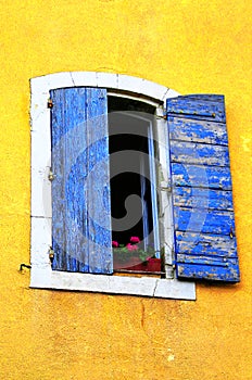 Old derelict window with blue wooden shutters with peeling paint on a yellow stucco facade.