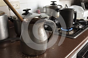 Old dirty cocoa cooking pot over kitchen furniture with gas stove on and others cooking pots at background.