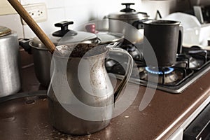 Old dirty cocoa cooking pot over kitchen furniture with gas stove on and others cooking pots at background.