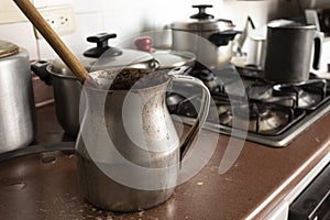 Old dirty cocoa cooking pot over kitchen furniture with gas stove on and others cooking pots at background.