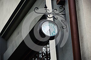 Old dirty round black outdoor clock with teal digits on black carved metal holder hanging on gray concrete wall