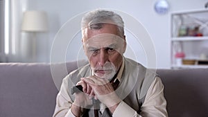 Old disabled man with walking stick sitting on couch, diseases at retirement age