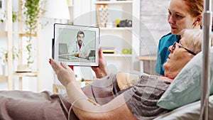 Old disabled woman lying in hospital bed having an online video call with a doctor