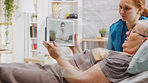 Old disabled woman lying in hospital bed having an online video call with a doctor