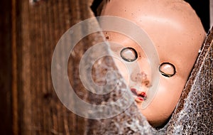 Old doll with eyes closed peering through a cobweb covered opening in a wooden door.