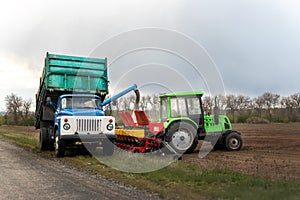 Old dumper medium truck near road loading agricultural seeder tractor filling planter with crops of wheat, corn or soybean for