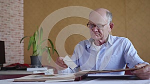 Old elderly lonely man in glasses for sight recalls happy moments of his youth leafing through photo album with photos