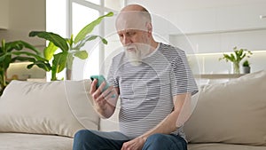 Old elderly pensioner communicates via smartphone via video chat with his family at distance. Grandpa is holding phone