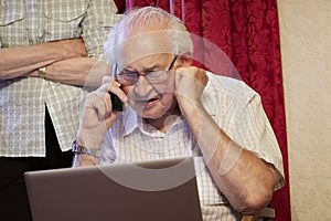 Old elderly senior man on phone at laptop computer at risk to cyber attack and online bank fraud