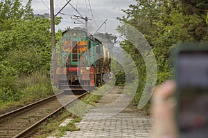 An old electric locomotive with a tank goes along a narrow-gauge railway past a tourist filming it on a smartphone photo