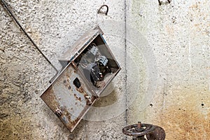an old electrical panel with switches or fuses torn off hangs crookedly on a bare concrete wall near a rusty iron valve