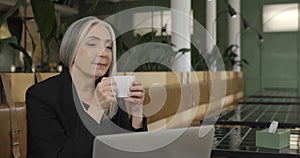 Old elegant businesswoman sitting at table and enjoing drinking coffee. Mature female enterpreneur working with laptop