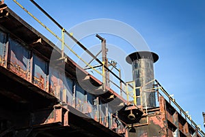 Old elevated train tracks heavy with rust and peeling paint, smoke stack behind as part of an abandoned industrial plant, blue sky