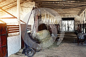 Old empty stables barn at horse farm with wooden beams , sulky cart and hay for feeding animals. Bright sunset sky